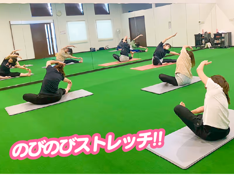 複数の人々が鏡のある部屋でヨガマットの上に座り、腕を伸ばして側面をストレッチしている。